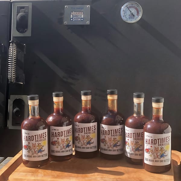 Six bottles of Hard Times BBQ sauce on a wooden tray in front of a large black smoker.