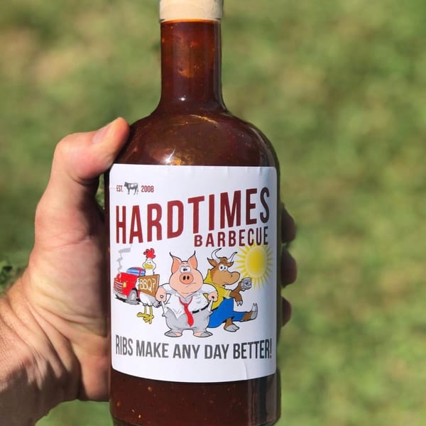 Hand holding a bottle of Hard Times Barbecue sauce with a cartoon label against a green background.