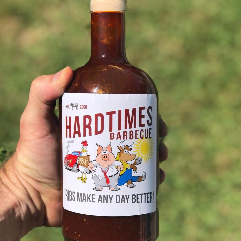 Hand holding a bottle of Hard Times Barbecue sauce with a cartoon label against a green background.