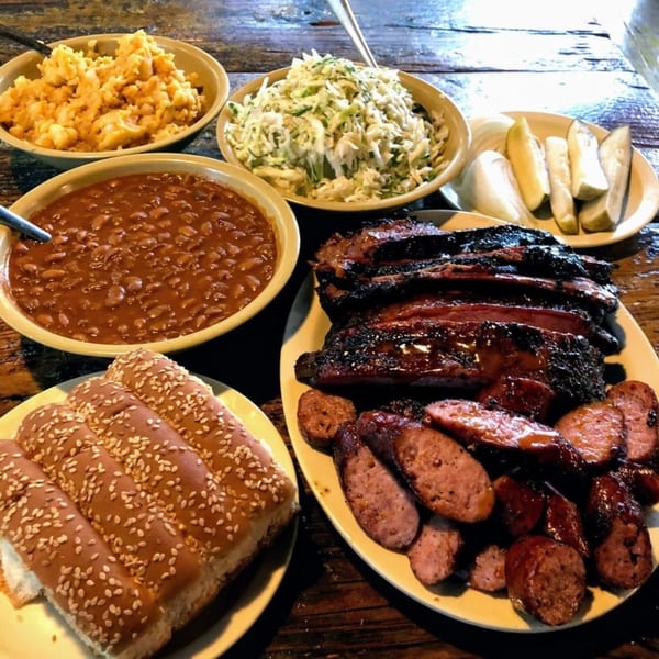 Hard Times BBQ spread with ribs, sliced sausage, baked beans, macaroni, coleslaw, pickles, and buns.