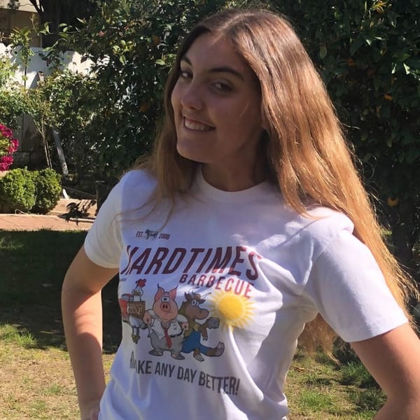 Smiling woman wearing a Hard Times BBQ t-shirt with cartoon pig, bull, and chicken characters.