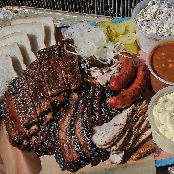 Hard Times BBQ platter with sliced brisket, sausage, turkey, coleslaw, baked beans, pickles, and onions.