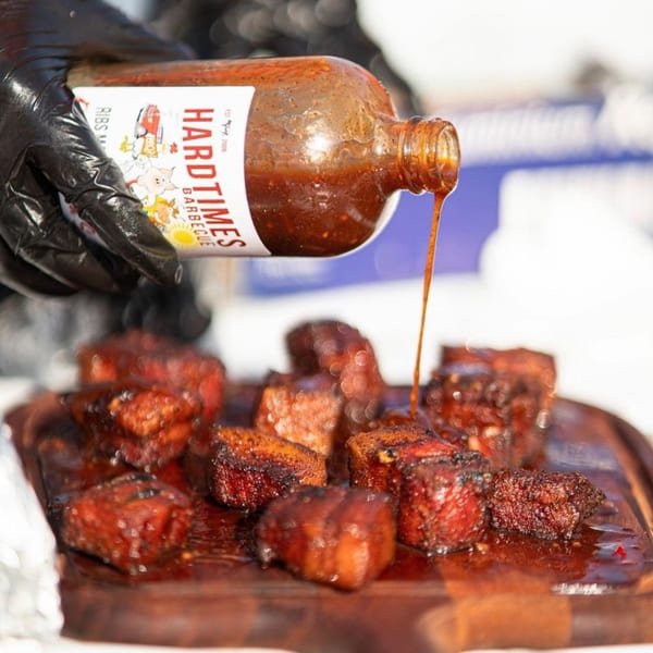 Gloved hand pouring Hard Times BBQ sauce over glistening burnt ends on a wooden board.
