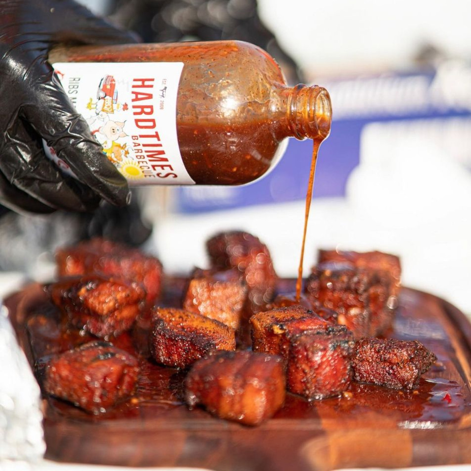 Gloved hand pouring Hard Times BBQ sauce over glistening burnt ends on a wooden board.