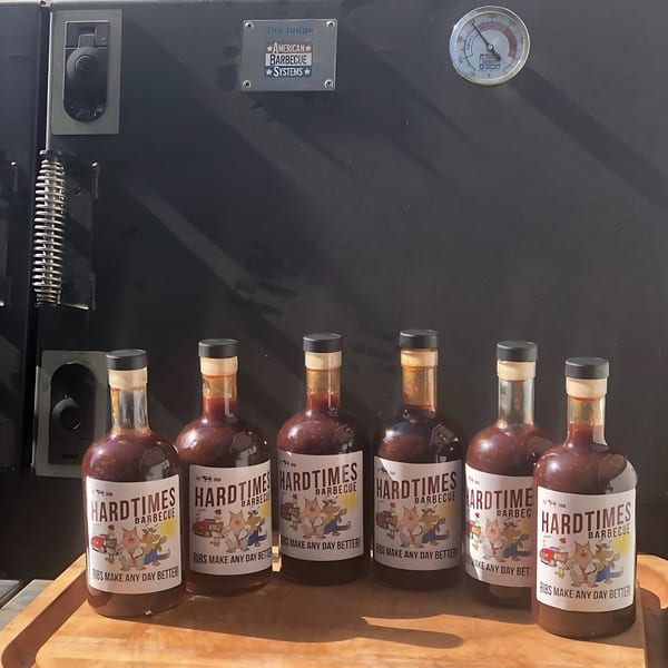 Six bottles of Hard Times BBQ sauce on a wooden tray in front of a large black smoker.