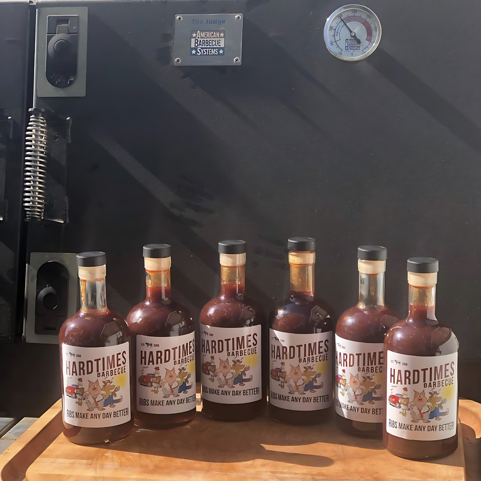 Six bottles of Hard Times BBQ sauce on a wooden tray in front of a large black smoker.