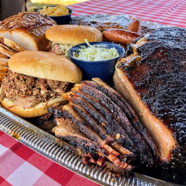 A large platter of Hard Times BBQ brisket, pulled pork sandwiches, and sides on a red checkered tablecloth.