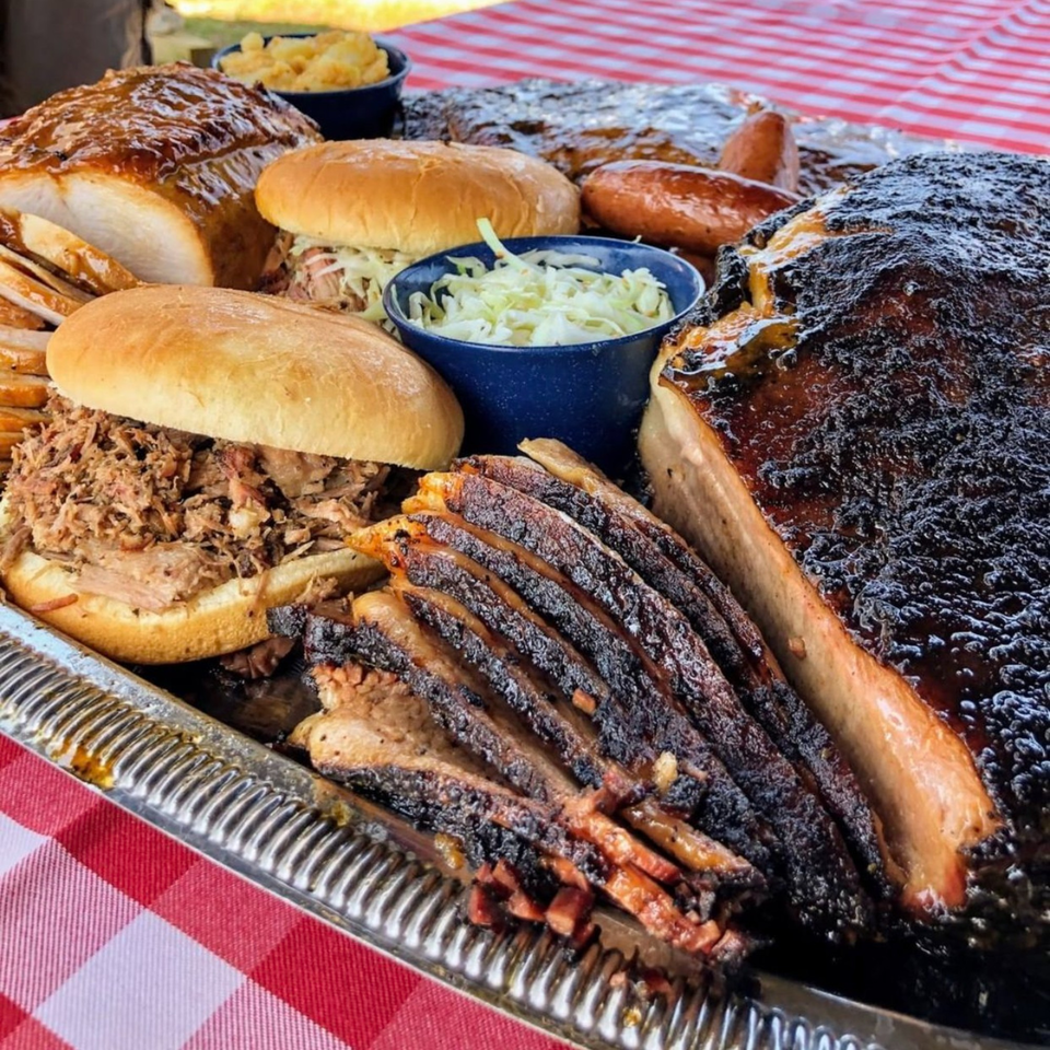 A large platter of Hard Times BBQ brisket, pulled pork sandwiches, and sides on a red checkered tablecloth.