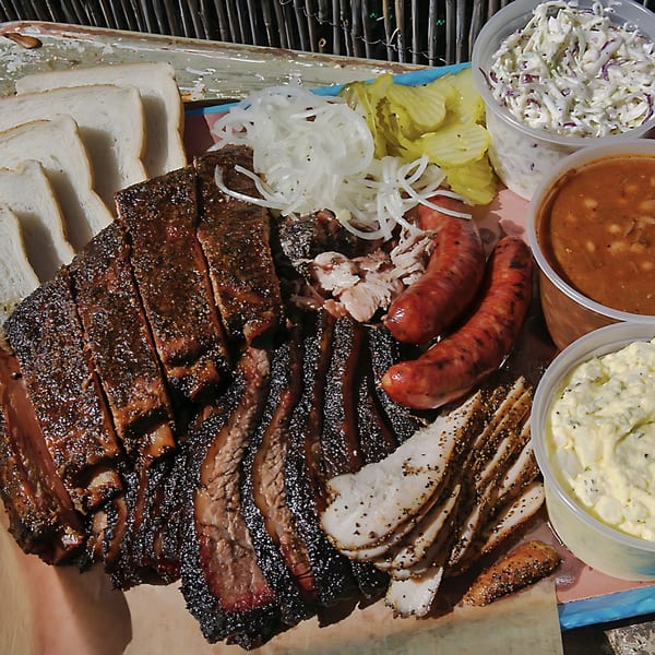 Hard Times BBQ platter with sliced brisket, sausage, chicken, white bread, onions, pickles, coleslaw, baked beans, and potato salad.